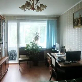 На фото: часть помещения жилой комнаты, большое окно, балконная дверь открыта, слева у стены - шкаф, справа у стены - письменный стол, стул, стены оклеены обоями, полы - ламинат, на потолке - люстра