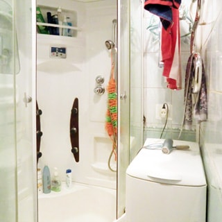 На фото: часть помещения санузла, установлена душевая кабина, справа у стены - стиральная машина с верхней загрузкой, над стиральной машиной на стене - полотенцесушитель, стены облицованы светлой керамической плиткой