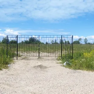 На фото: грунтовая дорога, отсыпанная гравием, упирается в металлические ворота перед въездом на поле, ворота на замке, забор отсутствует