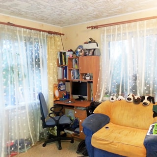 На фото: часть помещения жилой комнаты, комната угловая, два окна, между окнами в углу - компьютерный стол-стеллаж, офисное кресло, справа - мягкий диван