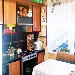 На фото: часть помещения кухни, окно, слева от окна вдоль стены встроенная кухонная мебель, 4-комфорочная газовая плита с духовкой, вытяжка, справа у окна - обеденный стол