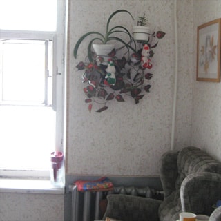 На фото: часть помещения жилой комнаты, окно, справа от окна на уровне подоконника - радиатор центрального отопления, в углу - мягкое кресло, на стене цветы в горшках, эстамп, стены оклеены обоями
