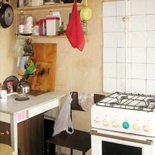 На фото: часть помещения кухни, газовая 4-комфорочная плита с духовкой, часть стены у плиты облицована белой керамической плиткой, слева от плиты - стул, кухонный столик, над ним - кухонные полки