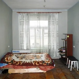 На фото: часть помещения жилой комнаты, одно трехстворчатое окно, слева у стены - диван-кровать, справа у стены - стеллаж и два табурета, полы - паркет