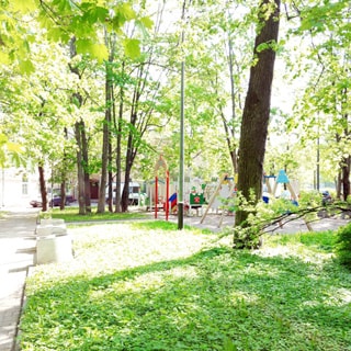 На летней фотографии: часть внутриквартальной придомовой территории, зеленые газоны, деревья, пешеходные дорожки, садовые скамейки, на заднем плане - детская площадка, за ней - жилой дом