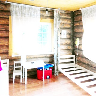 На фото: часть помещения детской комнаты, два окна, справа у окна - каркас детской кровати, слева у второго окна - детский стол со стулом и коробка с игрушками, стены бревенчатые, полы - ламинат, потолок - дощатый, на потолке - лампочка на проводе
