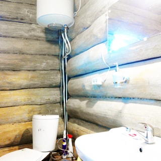 На фото: часть помещения совмещенного санузла, справа в углу установлен унитаз-компакт, над ним на стене - водогрейный электрокотел, справа - керамическая раковина со смесителем, над ней на стене - зеркало, стены - бревенчатые