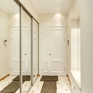 На фото: помещение прихожей, светлобежевые тона, слева - зеркальный шкаф-купе, справа - зеркало и тумба для обуви, в центре - входная дверь, домофон, полы - плитка, на потолке - точечные светильники
