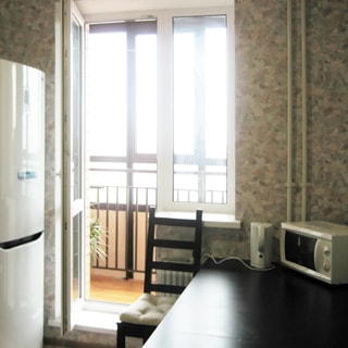 На фото: часть кухни, окно с балконной дверью, дверь на балкон открыта, балкон застеклен, слева от окна - двухкамерный холодильник, под окном - батарея центрального отопления, справа от окна - обеденный стол, на столе - электрический чайник и микроволновая печь, у стола - стул