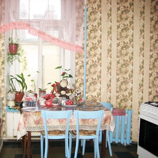 На фото: часть кухни, окно во двор, у окна - обеденный стол, справа от окна радиатор центрального отопления, еще правее у стены - газовая плита с духовкой, обои в цветочек, полы - линолеум.