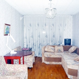 На фото: чистая комната в голубых тонах, мягкая мебель - два кресла слева и диван справа, слева у стены - стол, справа у окна - тумба с телевизором, светлые обои, полы - светлый паркет, на потолке - люстра
