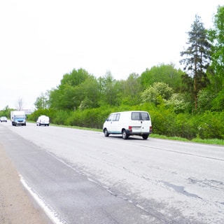 На фото: асфальтированная двухполосная дорога с отсыпаной обочиной, справа от дороги - кусты и лесополоса, по дороге двигаются одиночные легковые и грузовые автомобили