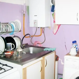На фото: часть помещения кухни, кухонная тумба с металлической мойкой, на мойке - смеситель, на тумбе и над ней - сушилка для посуды, справа от тумбы на стене - газовая колонка, правее - навесной кухонный шкаф, под ним - стиральная машина, стены - окрашены