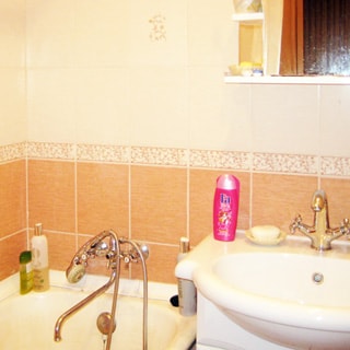 На фото: часть помещения ванной комнаты, керамическая раковина на стойке со смесителем, над ней полка и зеркало, слева от раковины - ванная со смесителем, стены облицованы керамической плиткой