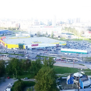 На фото: вид из окна, гипермаркет ОКей со своим открытым паркингом для автомобилей, проезжая часть с автомобилями и общественным транспортом, на дальнем плане торговый центр Континет и жилые кварталы