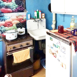 На фото: часть помещения кухни, прямо в углу - эмалированная мойка со смесителем, слева от мойки - четырехкомфорочная газовая плита с духовкой, справа у стены - холодильник, над ним - навесной кухонный шкаф, стены окрашены