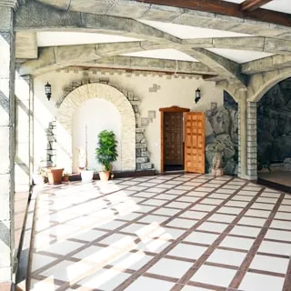 На фото: часть помещения просторного холла, слева падает свет от панорамных окон, помещение стилизовано под старинный замок, сводчатые каменные балки на потолке, колонны, арки, прямо на заднем плане - входная дверь, полы - плитка