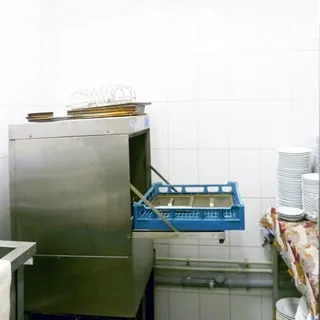 На фото: часть помещения для мойки посуды, стены в пол-высоты облицованы плиткой, прямо в углу - посудомоечная машина, справа у стены стол и стеллаж для чистой посуды