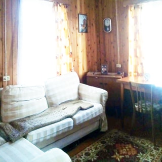 На фото: часть помещения жилой комнаты, два окна, между окон в углу - небольшой комод, справа от комода - письменный стол со стулом, слева от комода у окна - мягкий диван, стены отделаны вагонкой, на полу - ковер