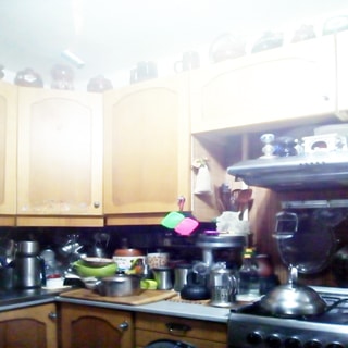 На фото: часть помещения кухни, кухонные столы-тумбы и навесные кухонные шкафы над ними, справа - четырехкомфорочная газовая плита, над ней - вытяжка, на столах - кухонная посуда, сверху на навесных полках - изделия из керамики