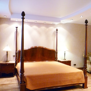 На фото: часть помещения жилой комнаты - спальни, прямо у стены - двуспальная кровать, слева и справа у изголовья - прикроватные тумбочки с настольными светильниками, правее у стены - кресло, на потолке - точечные светильники