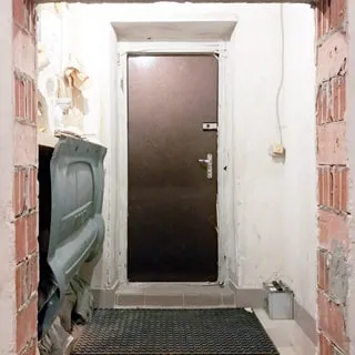 На фото: помещение входного тамбура, входная металлическая дверь с врезным и накладным замками, справа от двери на стене настенный выключатель света, полы - плитка, на полу - резиновый грязесборный коврик, вторая дверь тамбура не установлена, стены окрашены