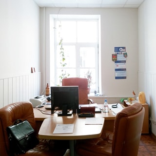 На фото: часть помещения офисного типа, одно окно, на окне - декоративные комнатные растения, перед окном - письменный стол с приставкой, за столом три больших кожаных офисных кресла, на столе - монитор компьютера и принтер, справа у окна в углу - тумбочка, на стене - календарь, полы - линолеум, стены окрашены, потолки - подвесные