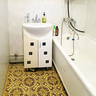 На фото: часть помещения ванной комнаты, справа вдоль стены полноразмерная ванна со своим смесителем, слева от нее на тумбе с дверцами - керамическая раковина со своим смесителем, полы и стены облицованы плиткой
