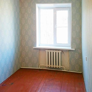 На фото: часть помещения жилой комнаты без мебели, одно окно, под окном - радиатор центрального отопления, полы - дощатые, стены оклеены обоями
