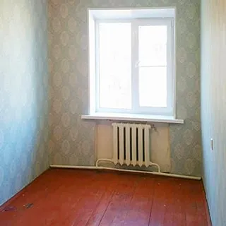 На фото: часть помещения жилой комнаты без мебели, одно окно, под окном - радиатор центрального отопления, полы - дощатые, стены оклеены обоями