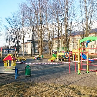 На фото: часть придомовой территории с большой детской площадкой и детским городком, вокруг площадки - деревья, на заднем плане жилые многокварирные кирпичные дома