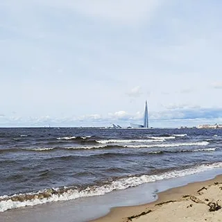 На летнем фото: пологий ровный песчаный берег большого водоема, на воде легкое волнение, на противоположном берегу - жилые дома и небоскреб Лахта центра