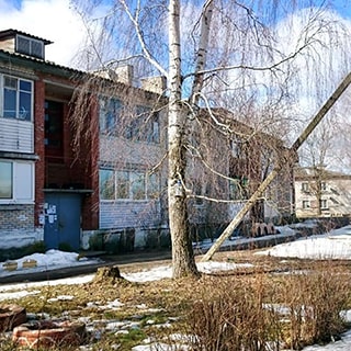 На фото: часть фасада двухэтажного кирпичного жилого дома, лоджии застеклены, кровля - шифер, на входе в парадную - металлическая дверь, перед фасадом - полисадник, асфальтированная дорожка, газон, деревья, кусты
