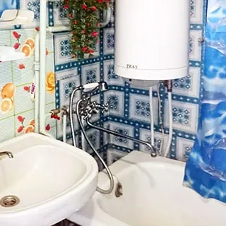 На фото: часть помещения ванной комнаты, справа вдоль стены полноразмерная ванна со своим смесителем и душем, над ней - электрический водогрей, слева от ванны - керамическая раковина со своим смесителем
