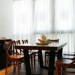 На фото: часть помещения кухни у большого панорамного окна от пола до потолка, перед окном - массивный дубовый обеденный стол на восемь человек, вокруг стола - деревянные стулья из массива дуба, стены - декоративная штукатурка, полы - керамическая плитка под дерево