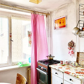 На фото: часть помещения кухни, одно большое двустворчатое окно во всю стену, под окном - радиатор батареи центрального отопления, справа у окна - четырехкомфорочная газовая плита с духовкой, справа от нее - кухонный стол-тумба, на плите - кастрюля, на столе - чашки, на потолке - лампа в абажуре, на стене над столом - кухонная полка с посудой, потолок и стены окрашены