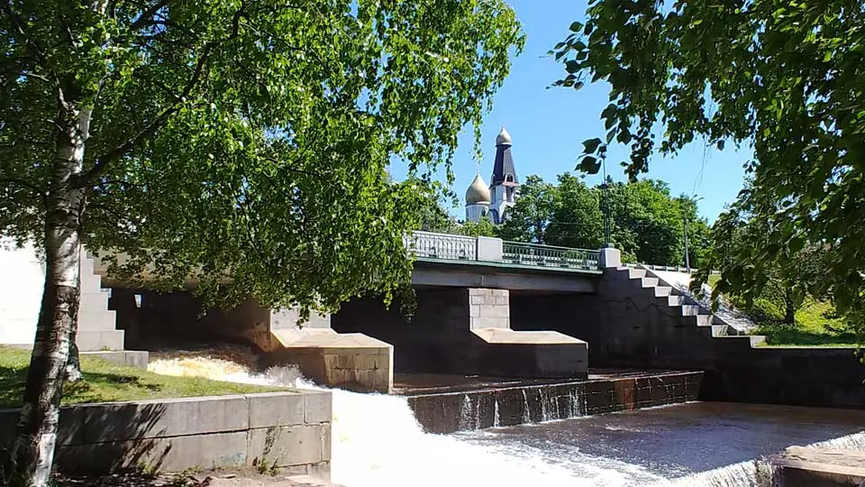 На летнем фото: плотина через водосливной канал, по плотине сверху - проезжая часть с тротуарами и перилами, много зелени, деревья, на дальнем плане - Петропавловская церковь Сестрорецка.