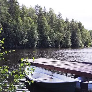 На летнем фото: небольшая лодочная станция с деревянным пирсом и тремя пришвартованными прогулочными лодками на веслах, водоем небольшого размера в окружении лесного массива, смешанный хвойно-лиственный состав деревьев