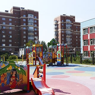 На летнем фото: оборудованная детским игровым городком детская площадка с полиуретановым покрытием, горками, лесенками, качелями и каруселями, на заднем фоне соседние жилые дома, справа за забором - здание и территория детского сада