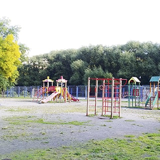 На летнем фото: оборудованная детским игровым городком детская площадка с горками, лесенками и качелями, за ней - огороженная сеткой спортивная площадка, за ней на заднем плане - деревья