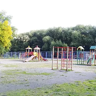 На летнем фото: оборудованная детским игровым городком детская площадка с горками, лесенками и качелями, за ней - огороженная сеткой спортивная площадка, за ней на заднем плане - деревья