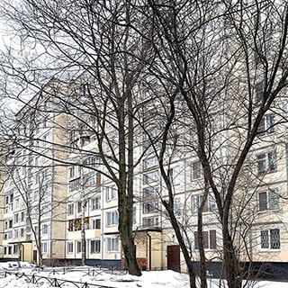 На зимнем фото: часть фасада девятиэтажного многоквартирного жилого панельного дома, большинство окон - стеклопакеты, часть квартир с балконами, входные группы с защитными козырьками, перед фасадом - огороженные газоны с деревьями