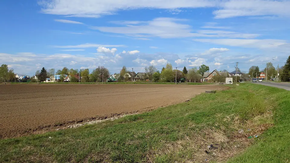На летнем фото: вспаханное поле, справа - асфальтированная автодорога, за полем - коттеджная застройка, между полем и домами - дорога, вдоль дороги - линия электропередач по железобетонным опорам.