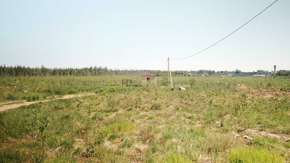 На летнем фото: земельный участок ровный, покрыт луговой растительностью, на среднем плане - небольшая постройка над артезианской скважиной, к ней подведена воздушная линия электропередачи и проложена грунтовая дорога, территория вокруг скважины огорожена забором из металлической сетки по металлическим столбам, на дальнем плане - лес