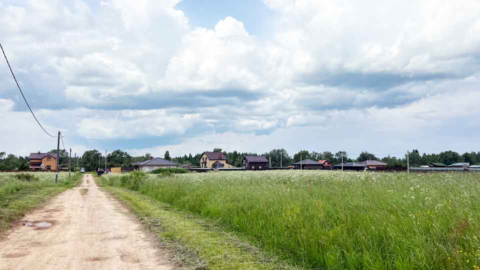 На летнем фото: проселочная грунтовая дорога, с правой и с левой стороны - луговые участки, вдоль левой стороны дороги воздушная линия электропередачи, на дальнем плане жилая малоэтажная застройка