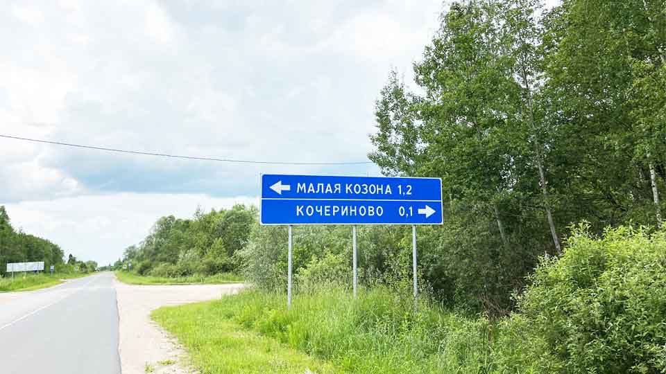 На летнем фото: асфальтовая дорога в окружении лиственных деревьев и кустарников, примыкание груновой дороги справа и слева с указателем населенных пунктов и расотяний до них