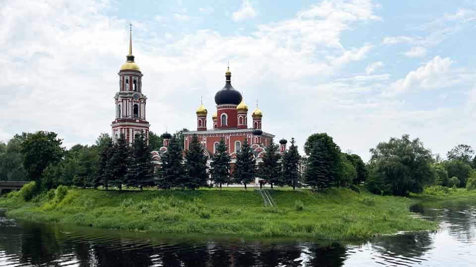 На летнем фото: на речном мысе на возвышении в окружении елей и лиственных деревьев - ансамбль Воскресенского собора с колокольней и позолоченными куполами