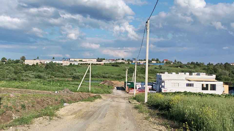 На летнем фото: проселочная грунтовая дорога, по правой стороне ЛЭП по бетонным столбам, справа и слева от дороги - полевой ландшафт, на дальнем плане и впереди справа жилые и хозяйственные постройки