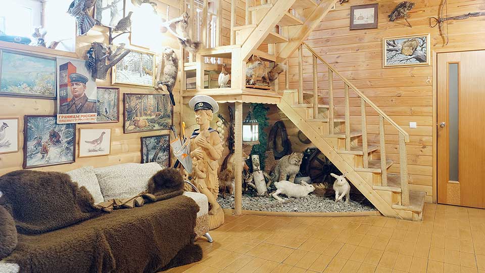 На фото: часть просторного нежилого помещения - прихожей, полы - плитка, стены отделаны строганой доской, в левом дальнем углу - деревянная двухпролетная лестница на второй этаж, слева у стены - мягкий диван, в дальней стене - закрытая дверь в соседнее помещение