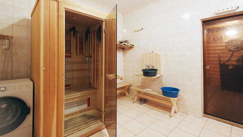На фото: часть просторного нежилого помещения - прачечной, полы и стены в полную высоту - плитка, потолки окрашены, прямо у стены стиральная машина с фронтальной загрузкой, ифракрасная сауна, справа в стене - стеклянная дверь в сауну